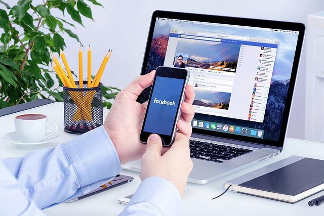 Person holding a smartphone with a laptop in the background displaying a Facebook page. 